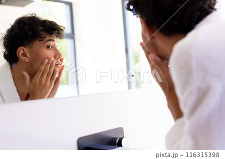 Looking in mirror, young man touching face and examining skin in bathroom 116315398