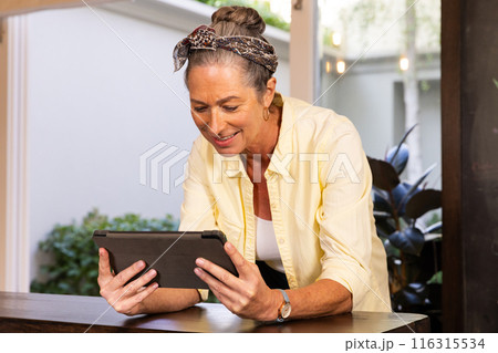 Smiling senior woman using tablet while standing at home, enjoying technology 116315534