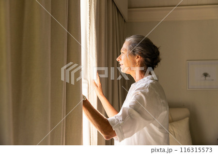 Looking out window, mature woman in white shirt enjoying peaceful moment at home, copy space 116315573
