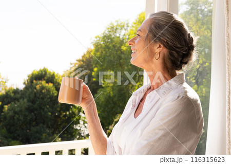 Smiling mature woman holding coffee mug and looking out window at home, copy space Smiling mature woman holding coffee mug and looking out window at home, copy space 116315623
