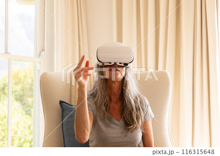 Using VR headset, mature woman sitting on chair interacting with virtual environment Using VR headset, mature woman sitting on chair interacting with virtual environment 116315648