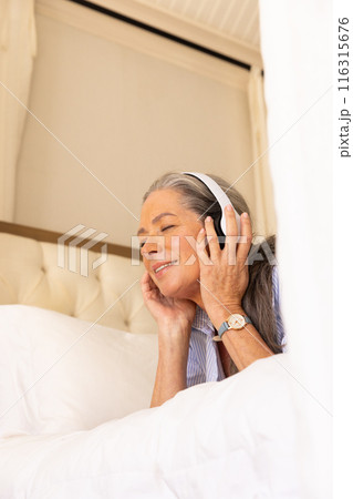 Listening to music, mature woman relaxing on bed with headphones on 116315676