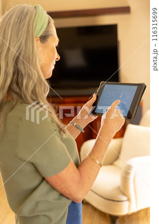 Using tablet, mature woman managing smart home devices in living room 116315699