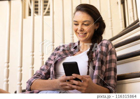 Smiling young woman using smartphone while sitting on stairs at home 116315864