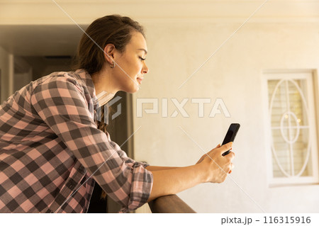 Using smartphone, young woman leaning on railing and smiling at home Using smartphone, young woman leaning on railing and smiling at home 116315916