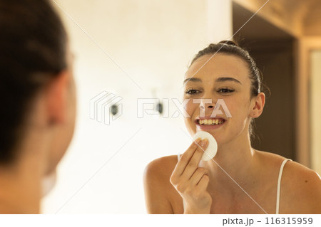 Smiling woman using cotton pad for skincare routine in bathroom mirror 116315959