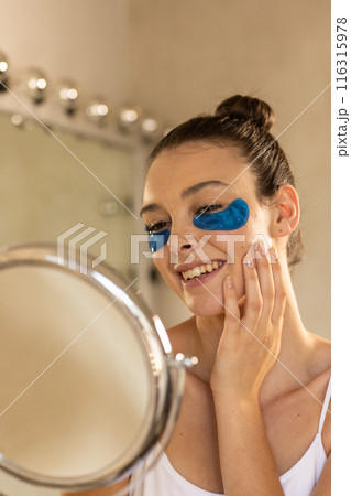 Applying under-eye patches, woman smiling and looking in mirror at home 116315978