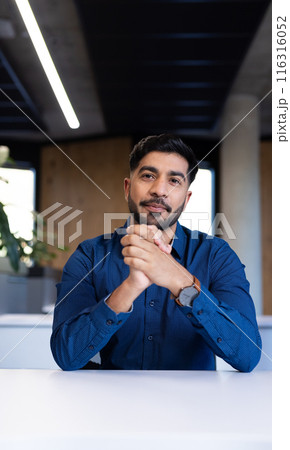 Businessman in blue shirt sitting at desk, confidently looking at camera, copy space 116316052