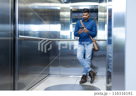 Using smartphone, man standing in elevator, wearing casual business attire, copy space Using smartphone, man standing in elevator, wearing casual business attire, copy space 116316191