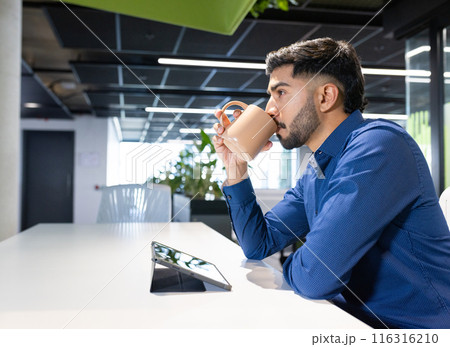 Using tablet, man drinking coffee in modern office workspace, copy space 116316210