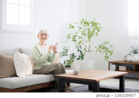 ソファーに座ってスマートフォン(スマホ)を持つ笑顔のシニア女性　カメラ目線　タップや操作する 広角 116320957