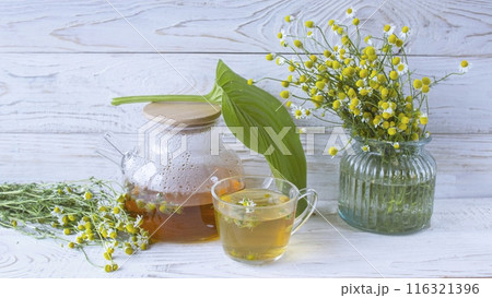 Herbal medicinal tea with chamomile flowers in a glass teapot, a bouquet of fresh flowers, on a wooden light background. Herbal medicinal tea with chamomile flowers in a glass teapot, a bouquet of fresh flowers, on a wooden light background. 116321396