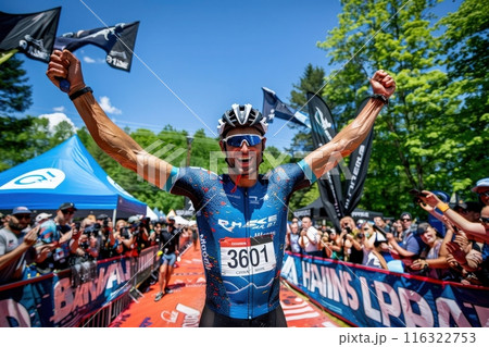 A cyclist raises his arms in victory as he crosses the finish line of a race, surrounded by cheering spectators. 116322753