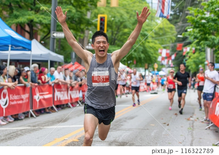A male runner in a grey tank top crosses the finish line in a race, celebrating with raised arms. Other runners are in the background. 116322789