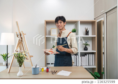 Young Artist Painting in Modern Studio with Canvas and Art Supplies, Smiling at Camera, Surrounded by Plants and Bookshelves Young Artist Painting in Modern Studio with Canvas and Art Supplies, Smiling at Camera, Surrounded by Plants and Bookshelves 116325599