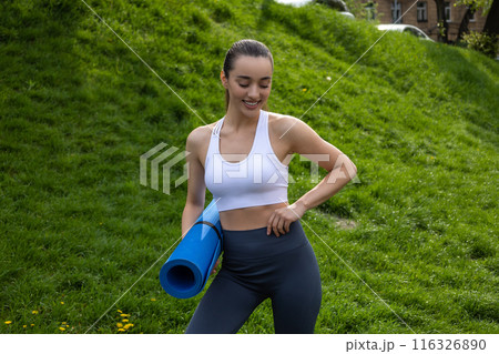 Happy young girl holding yoga mat smiling while preparing for workout in park 116326890