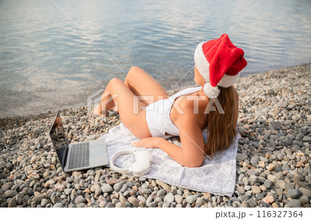 A woman is laying on the beach with a laptop and headphones on her head. She is wearing a red Santa hat. 116327364