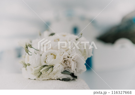 A bouquet of white flowers sits on a ledge next to a blue building. The flowers are arranged in a vase and are the main focus of the image. The blue building in the background adds a sense of calm. 116327668