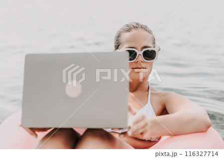 A woman is sitting in a pink inflatable raft on a lake, holding a laptop. Concept of relaxation and leisure, as the woman enjoys her time in the water while working on her laptop. A woman is sitting in a pink inflatable raft on a lake, holding a laptop. Concept of relaxation and leisure, as the woman enjoys her time in the water while working on her laptop. 116327714