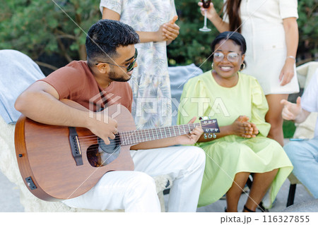 Multiracial group of people, indian man playing guitar and friends eating dinner and drinking wine during party in the picnic 116327855