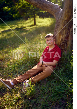 preteen boy in a red t-shirt is sad near a withered tree in the park 116328669