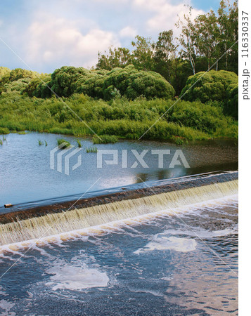 Serene natural scene with small waterfall, calm body of water, lush green vegetation, partly cloudy sky. Peaceful, scenic setting in forested area. 116330337