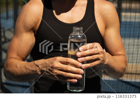 Athletes man holding glass bottle of water on outdoor sports ground. Concept water balance for hot season for active workout. Close up. 116331375