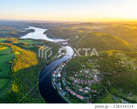 A sunset aerial shot of Orlik Reservoir in Czechia shows a winding river, hills, and a quaint town in the valley with vibrant blue and golden skies. A sunset aerial shot of Orlik Reservoir in Czechia shows a winding river, hills, and a quaint town in the valley with vibrant blue and golden skies. 116334314