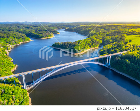 An aerial view of the Zdakov Bridge spanning the Vltava River in the Czech Republic. The bridge arches gracefully over the water, surrounded by lush green forests. 116334317