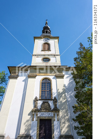 A low-angle view of Saint Lawrence Church in Potstejn, Czechia, showcasing the churchs facade, bell tower, and a sign reading Charitas. 116334371