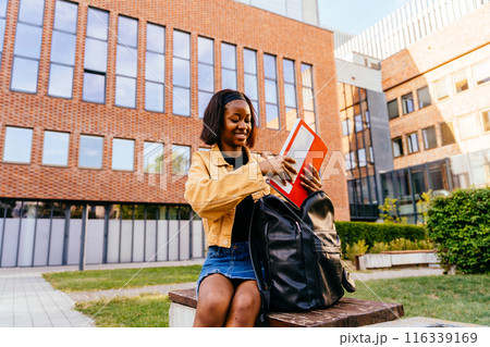 A young woman is sitting on a bench with backpack and exercise book. She is smiling and she is enjoying her time. Concept of relaxation and leisure 116339169