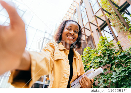 African woman student smiling to camera, making selfie photo and holding a stack of books. Concept of happiness and accomplishment, as the woman is proud of her studies or her current situation. 116339174