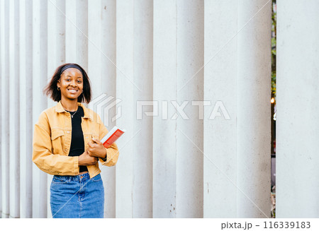 Portrait of black female student at campus looking at camera with modern building on background. Education, school and people concept. Portrait of black female student at campus looking at camera with modern building on background. Education, school and people concept. 116339183