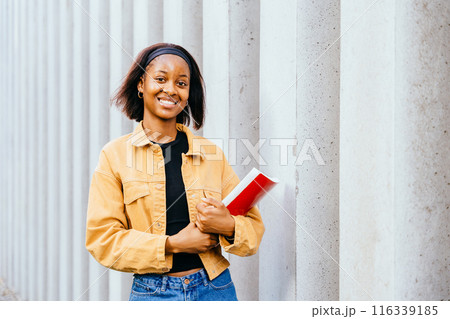 A young happy smiling African woman student university person ready for learning, goals or education. Female wearing a yellow jacket and a black shirt is holding a red notebook. Education, school and A young happy smiling African woman student university person ready for learning, goals or education. Female wearing a yellow jacket and a black shirt is holding a red notebook. Education, school and 116339185