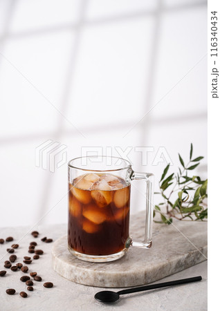 Iced coffee in a cup with ice cubes and grains on a light marble background with morning shadows. Iced coffee in a cup with ice cubes and grains on a light marble background with morning shadows. 116340434