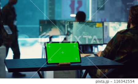 Tablet with greenscreen display running in a command post base of operations, military personnel ensuring national safety in control room. Soldiers and commanders use radar surveillance. Camera A. 116342554