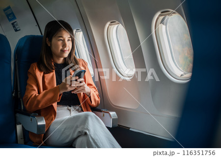 Woman seated on airplane, using smartphone, wearing orange blazer and white pants, looking out window, in-flight travel. 116351756