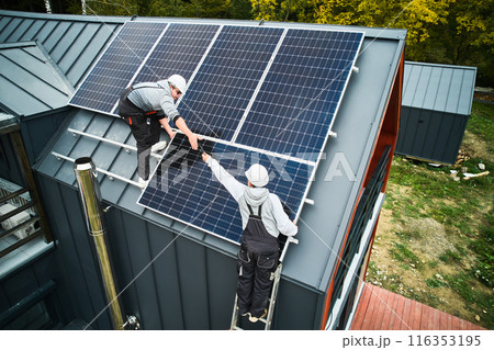 Builders building photovoltaic solar module station on roof of house. Men electricians in helmets installing solar panel system outdoors. Concept of alternative and renewable energy. Aerial view. Builders building photovoltaic solar module station on roof of house. Men electricians in helmets installing solar panel system outdoors. Concept of alternative and renewable energy. Aerial view. 116353195