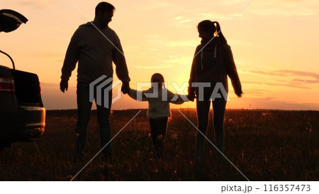 Family holding hands at sunset, silhouettes near car. Joint recreation in nature. Mother father daughter in orange setting sun in field. Family picnic road trip, parents kid enjoy each other's company 116357473
