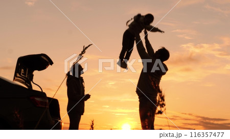 Silhouette of father tossing child in air. Family traveling by car having fun in nature laughing smiling enjoying life. Happy moments together. Loving dad with daughter and wife at weekend travel trip 116357477