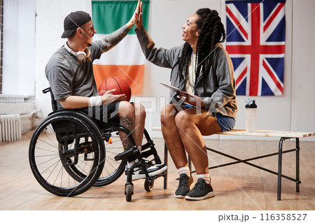 Side view portrait of young sportsman with disability high five with smiling girl in indoor basketball court copy space Side view portrait of young sportsman with disability high five with smiling girl in indoor basketball court copy space 116358527