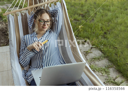 young adult girl in a hammock works on a computer outside the house and smokes an electronic cigarette young adult girl in a hammock works on a computer outside the house and smokes an electronic cigarette 116358566