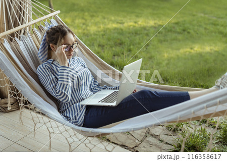 Young beautiful woman lies in a hammock with a laptop in glasses outside at home Young beautiful woman lies in a hammock with a laptop in glasses outside at home 116358617
