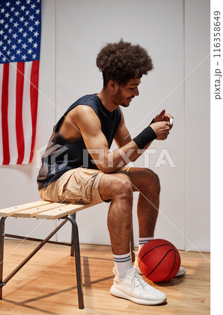 Full length side view portrait of Black young man as basketball player preparing for match sitting on bench in locker room with USA flag in background 116358649