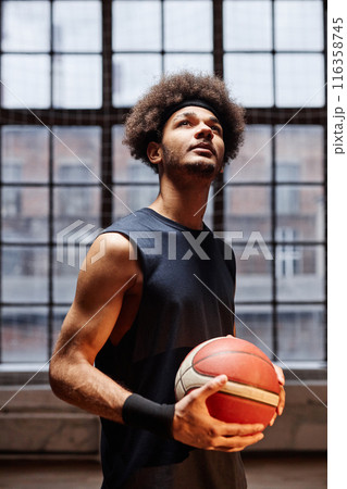 Vertical portrait of young African American man as a basketball player holding ball and looking up in dramatic lighting against window 116358745