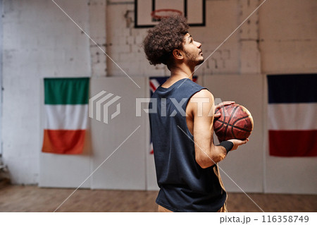 Back view of young African American teenage boy as a basketball player holding ball and looking up in gym with contour light 116358749