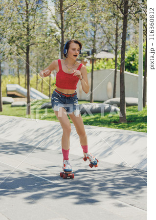 Radiant skater girl in vintage red crop top and denim shorts, listening music in headphones and enjoying a sunny day outside. 116360812
