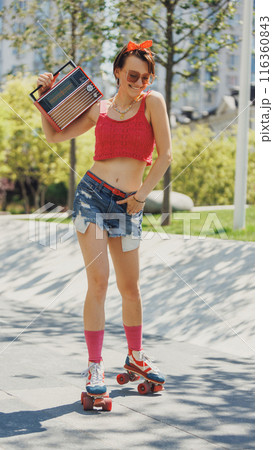 Young woman in vintage casual summer wear, roller skating with vintage radio in her hands, her face lit up with joyful smile. 116360843