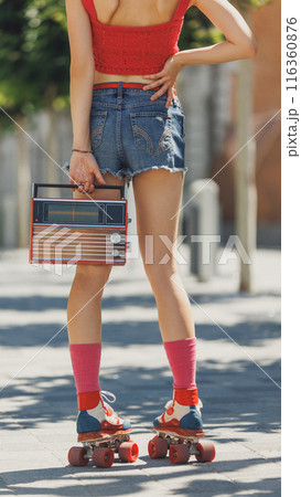 Cropped photo of young woman in red crop top and denim shorts roller skating with vintage radio in hand, beaming with joy on sunny day. 116360876