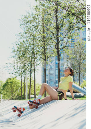 Cheerful skater girl taking break on sunny day, seated on ramp with her retro roller skates on, dressed in vibrant summer attire. 116360901